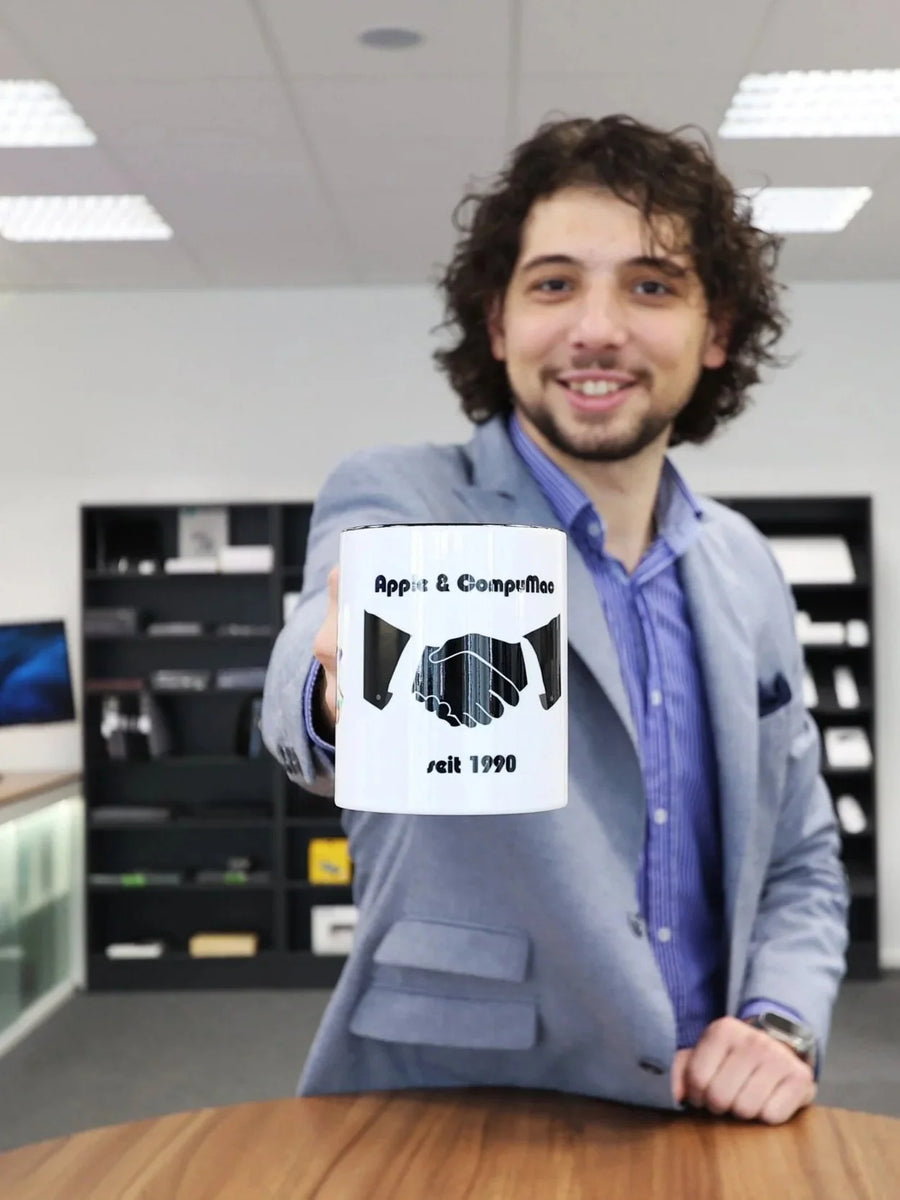 A person holds a mug printed with “Apple & CompuMac seit 1990” up to the camera.